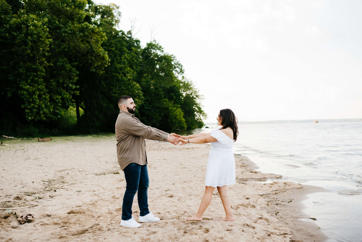 man and woman hold hands and dance together on the beach at woodbridge lake on a cloudy day with a forrest on the edge of the beach  photographed by Virginia wedding photographer