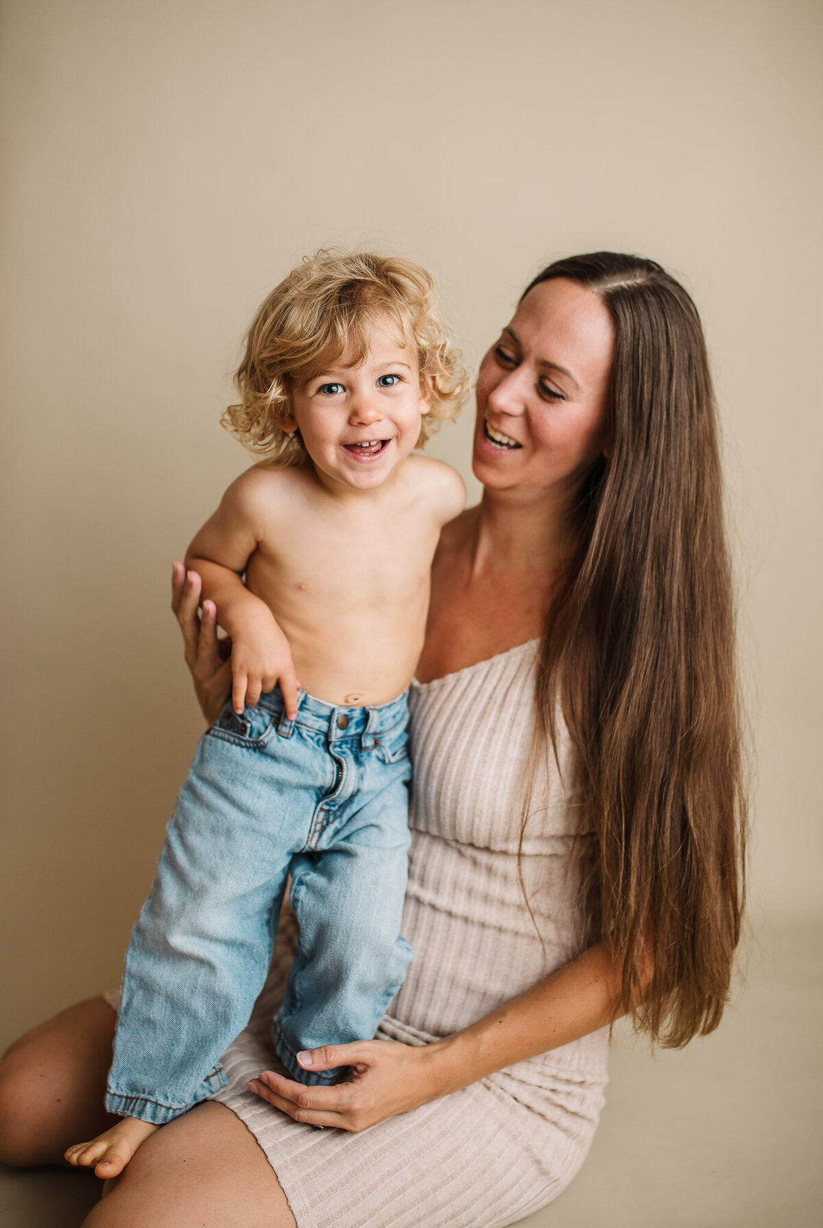 Toddler boy standing on mom's lap wearing only denim jeans, mom is looking down laughing at him he is looking and smiling at camera