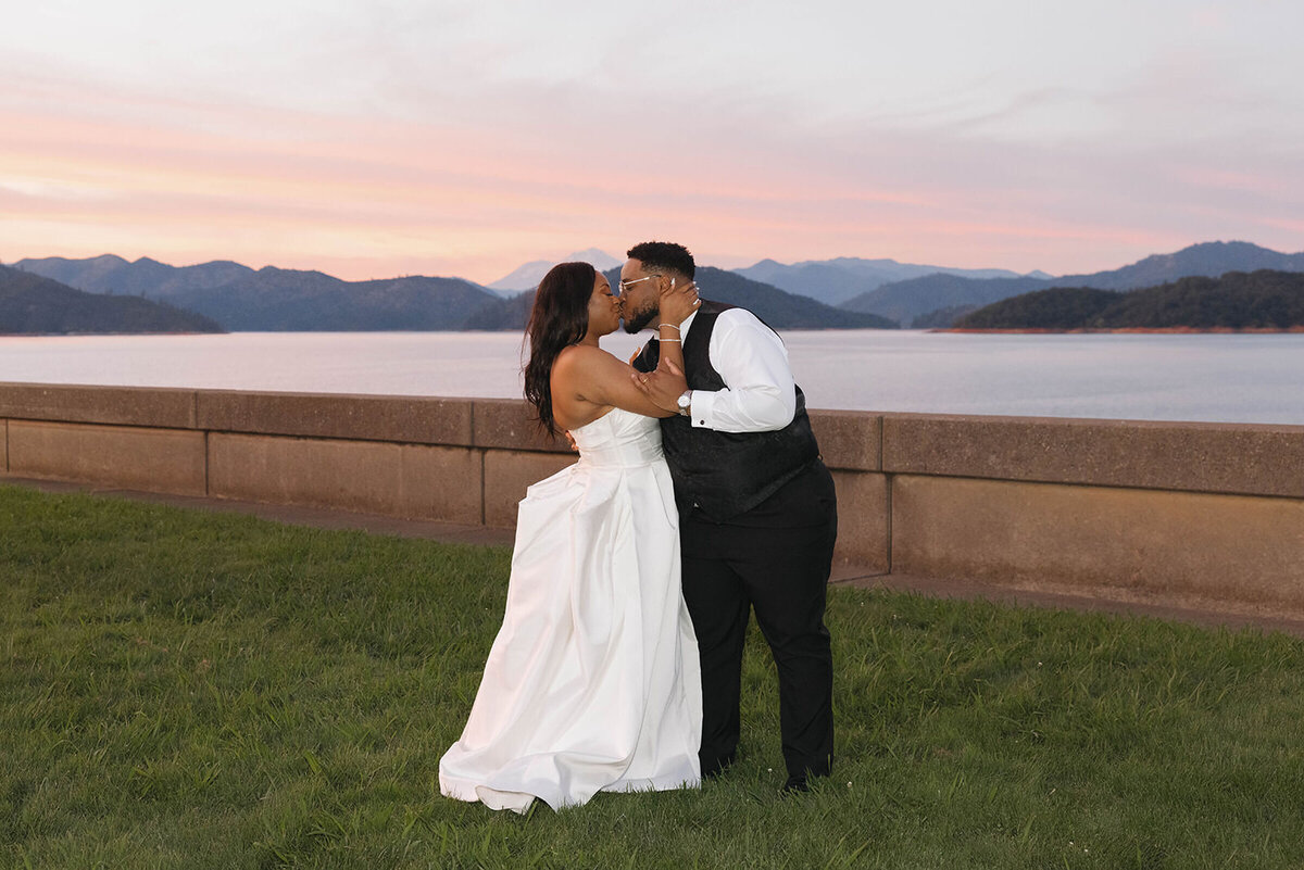 Groom dipping his bride for a kiss under a pink sunset sky by the lake