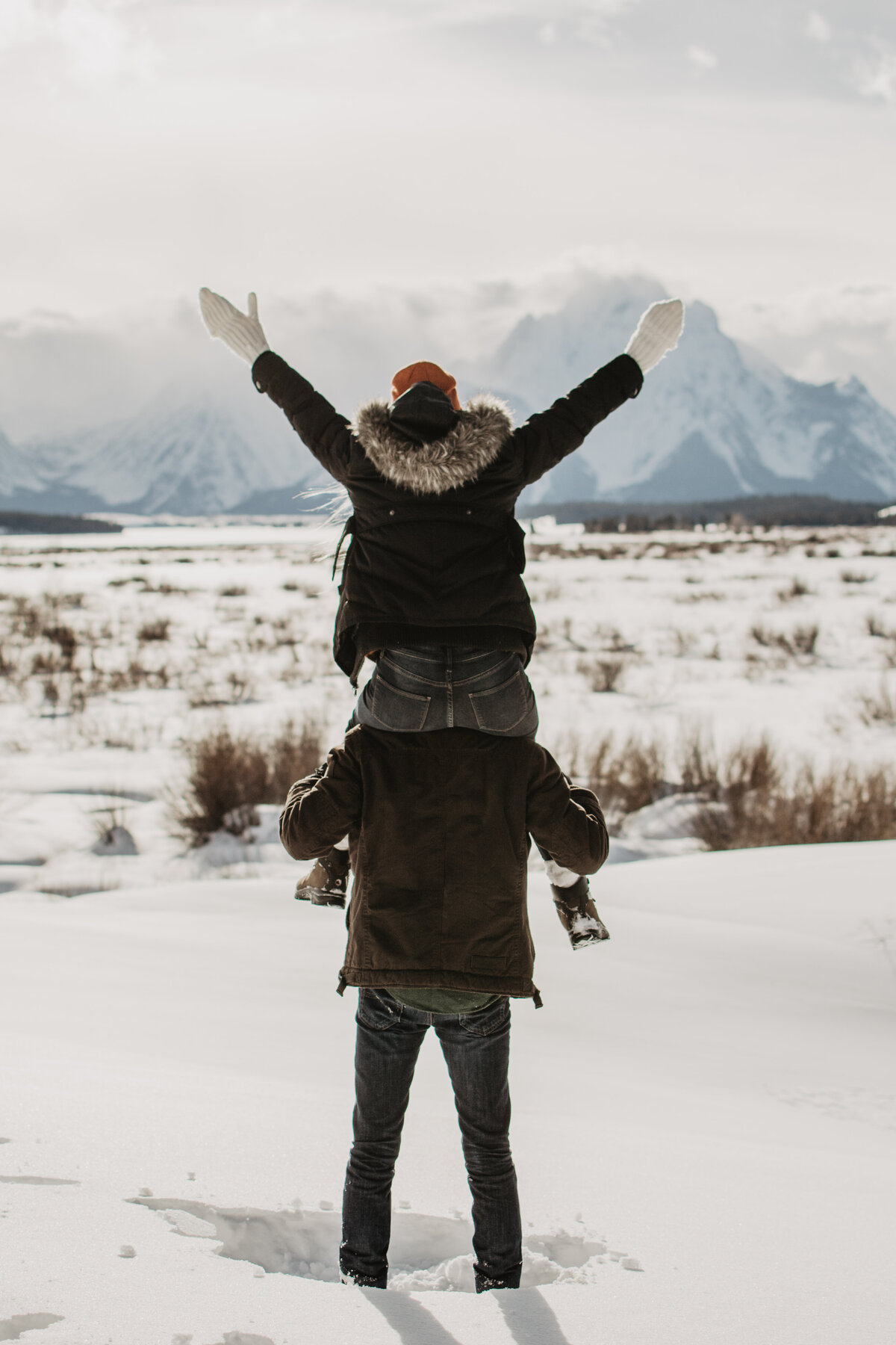 jackson hole photographers photographs woman on mans shoulder with her arms up in the air as they are faced away from the camera and looking at the Grand Tetons for their winter engagement session