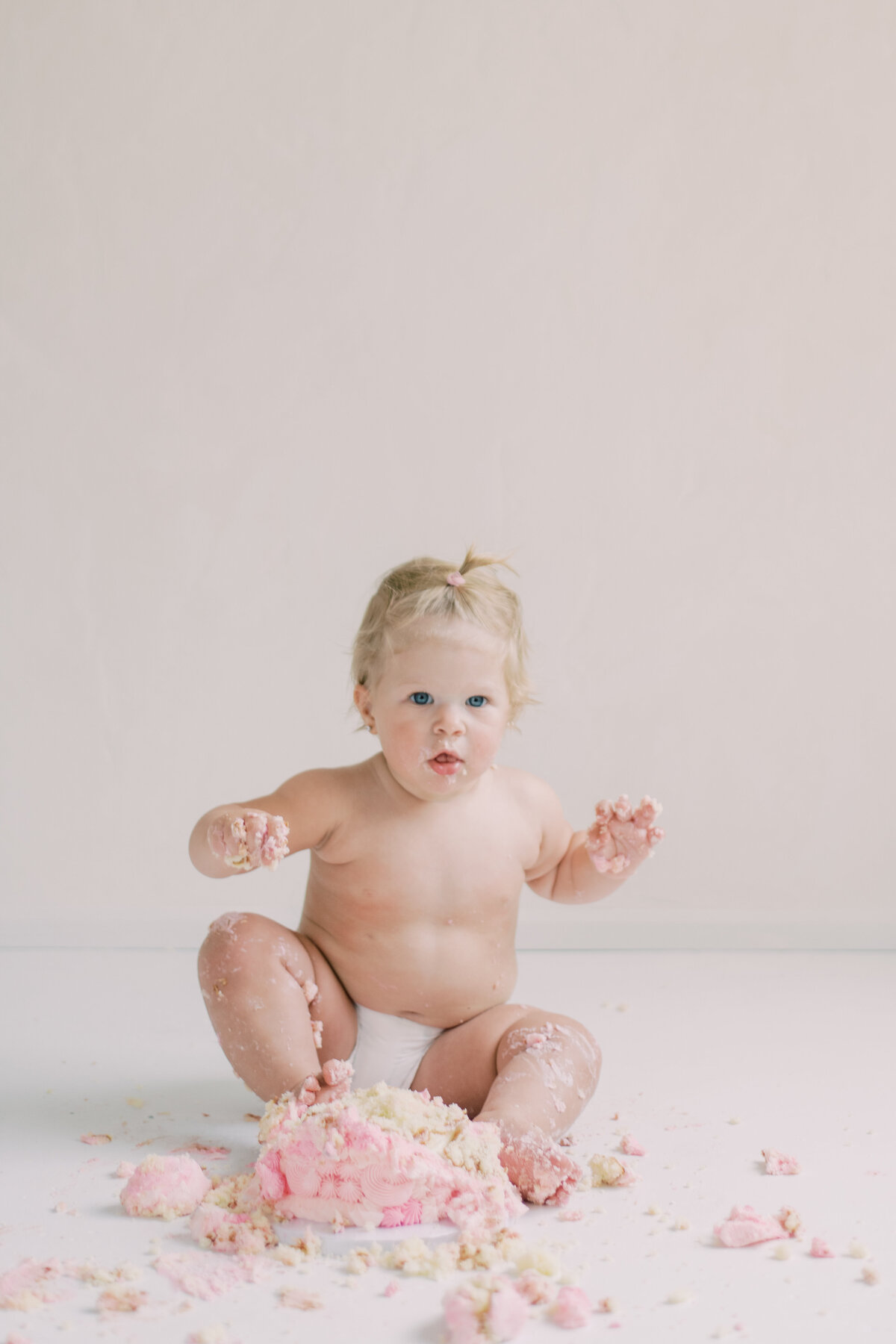 Baby girl in crown seated against white backdrop, mid-laugh, surrounded by soft pastel cake