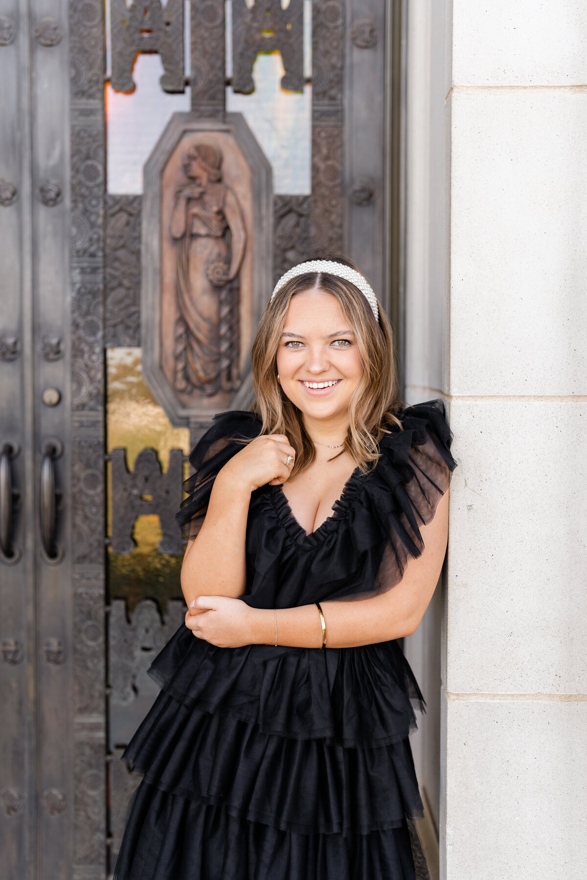 Texas A&M senior girl wearing black fluffy dress while leaning against Administration Building while holding elbow and touching hair wearing pearl headband