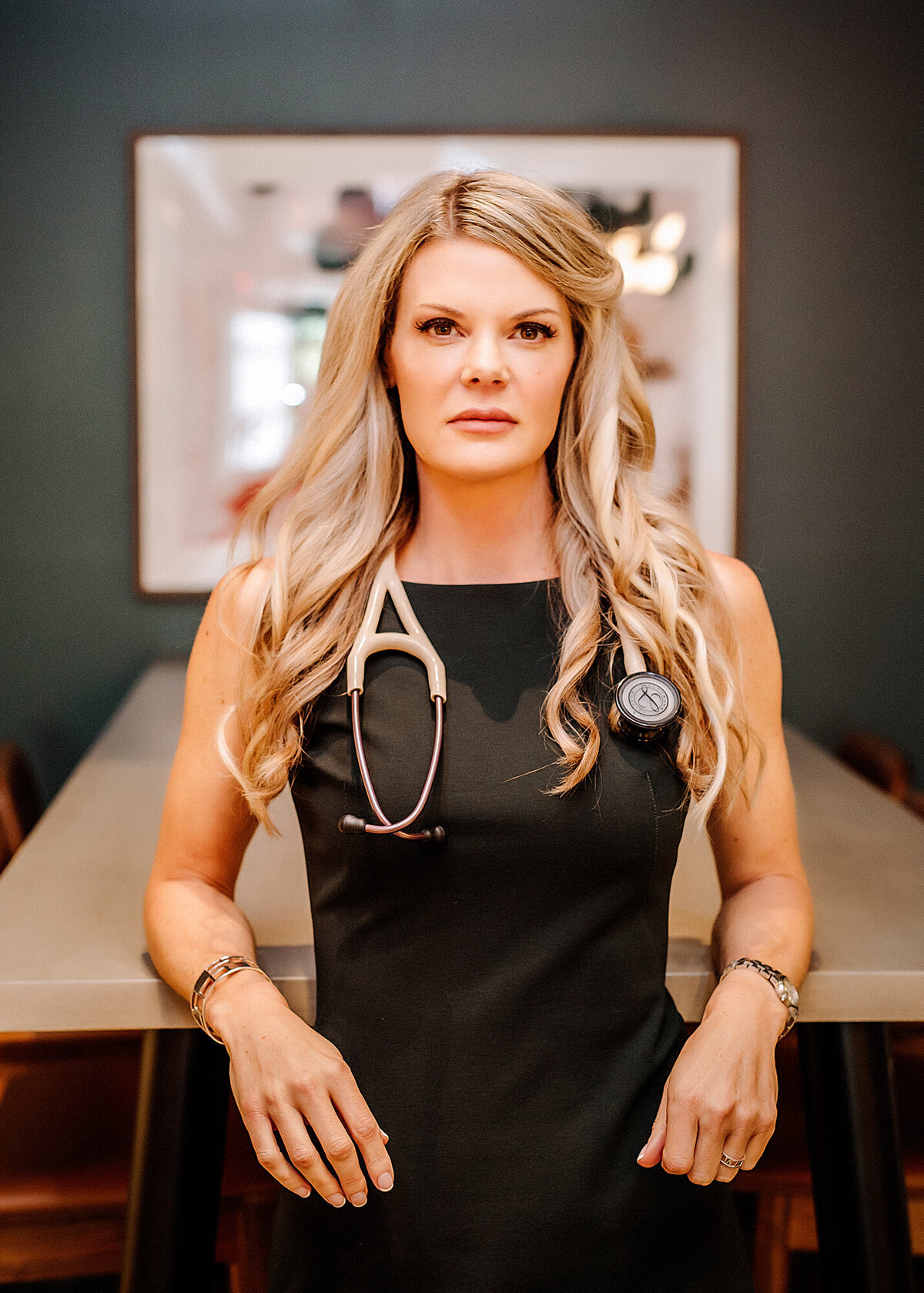 A Female Nurse Practitioner wearing a black dress, standing up against a table with artwork behind her, in a power pose for a South Shore MA branding photographer