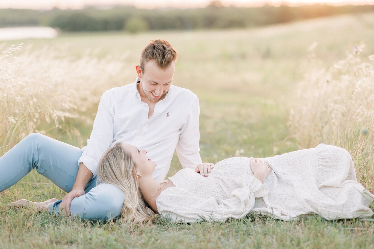 Husband with his expecting wife relaxing in a field at sunset while mama holds baby bump and they are smiling at one another