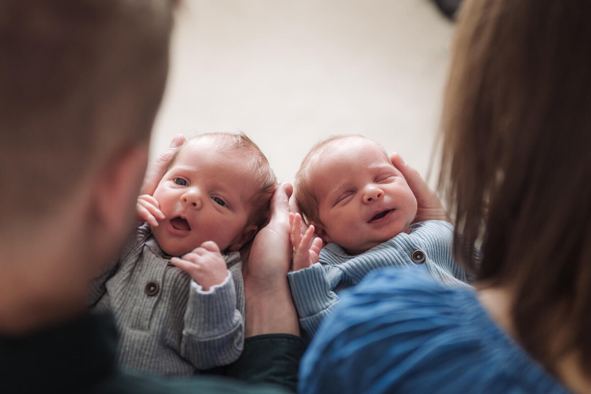 Twin newborns posing for newborn photos looking through parents heads from behind