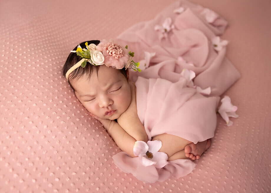 baby girl posed bum up on a soft pink backdrop with a pink chiffon drape and flowers draped over her.