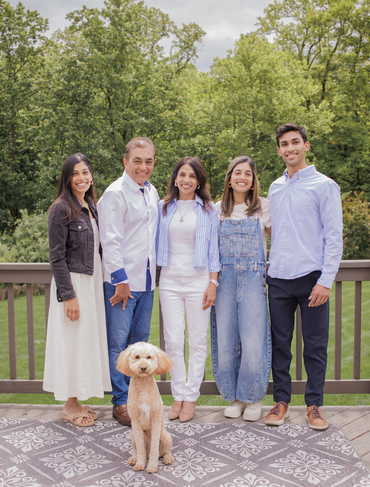 Smiling family with their dog posing together outdoors on a deck surrounded by trees during a summer family photo session