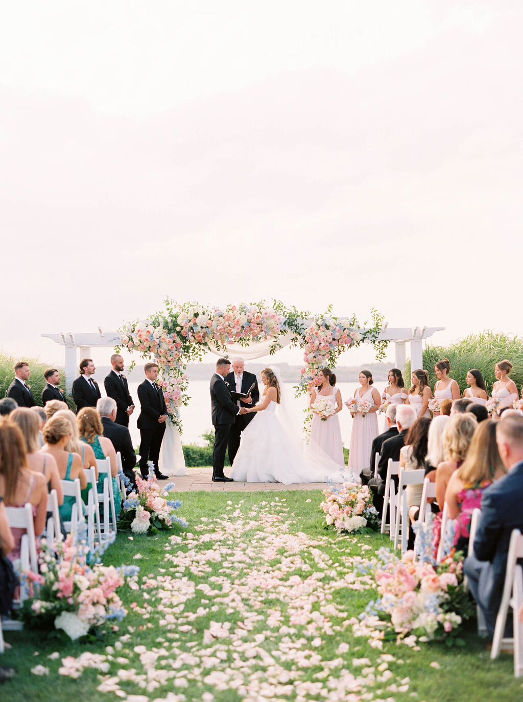 Newport Wedding Photographer | Outdoor wedding ceremony with a bride and groom under a floral arch. Guests are seated on white chairs, surrounded by greenery and pastel flowers.