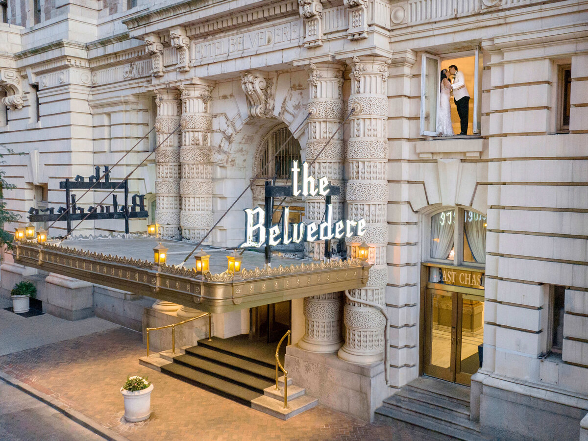 A grand hotel entrance with The Belvedere sign is adorned with ornate columns and lights. A couple in wedding attire stands at an upper window, sharing a moment. The street below is cobblestone with potted plants.