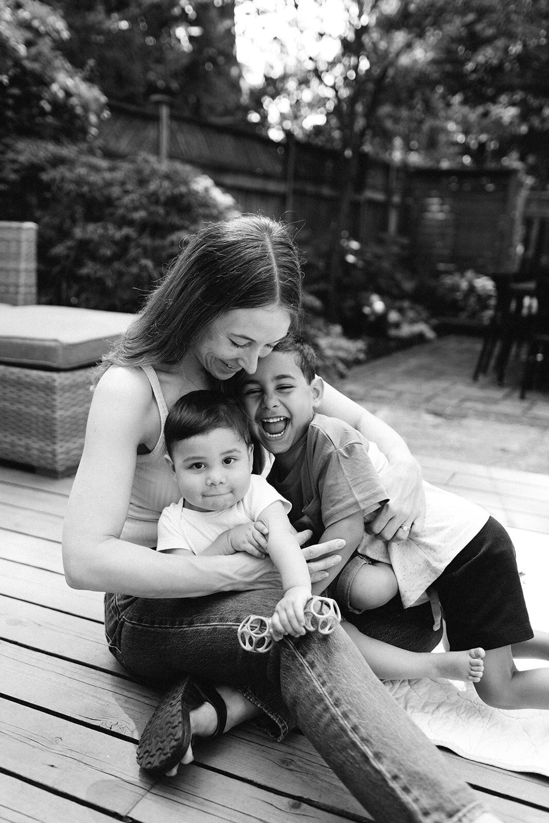 A woman sits on a wooden deck, hugging two children for their Toronto in home family photography, captured by Jennifer van Son. One child holds a small object. All are smiling. The background has outdoor furniture and greenery in this classic black and white image.