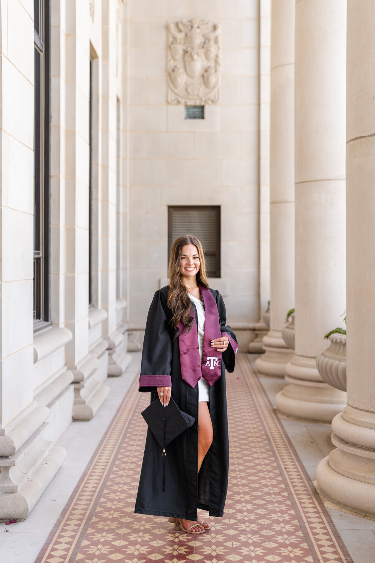 Texas A&M senior girl standing with hand on stole while wearing gown, stole and holding cap by her side in middle of columns of Administration Building
