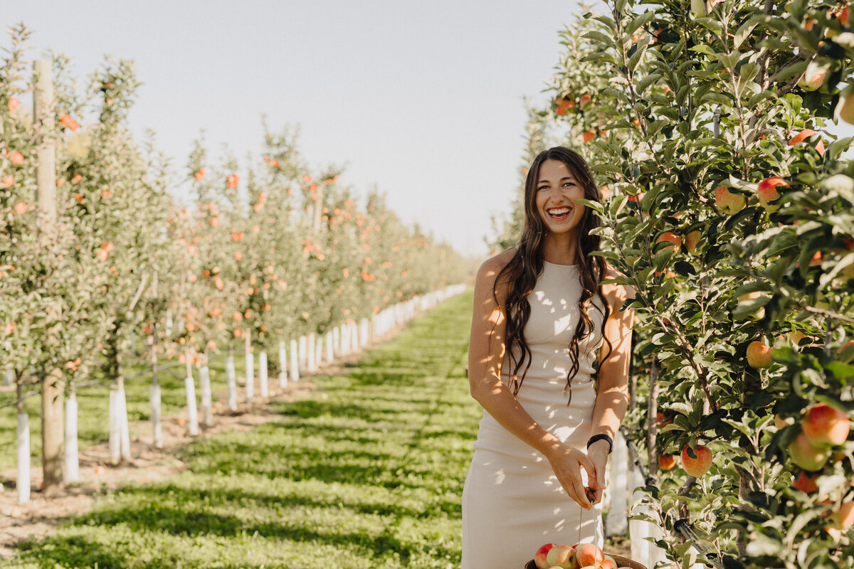 A soon-to-be bride in a white dress smiling and posing amongst the trees in the orchard.