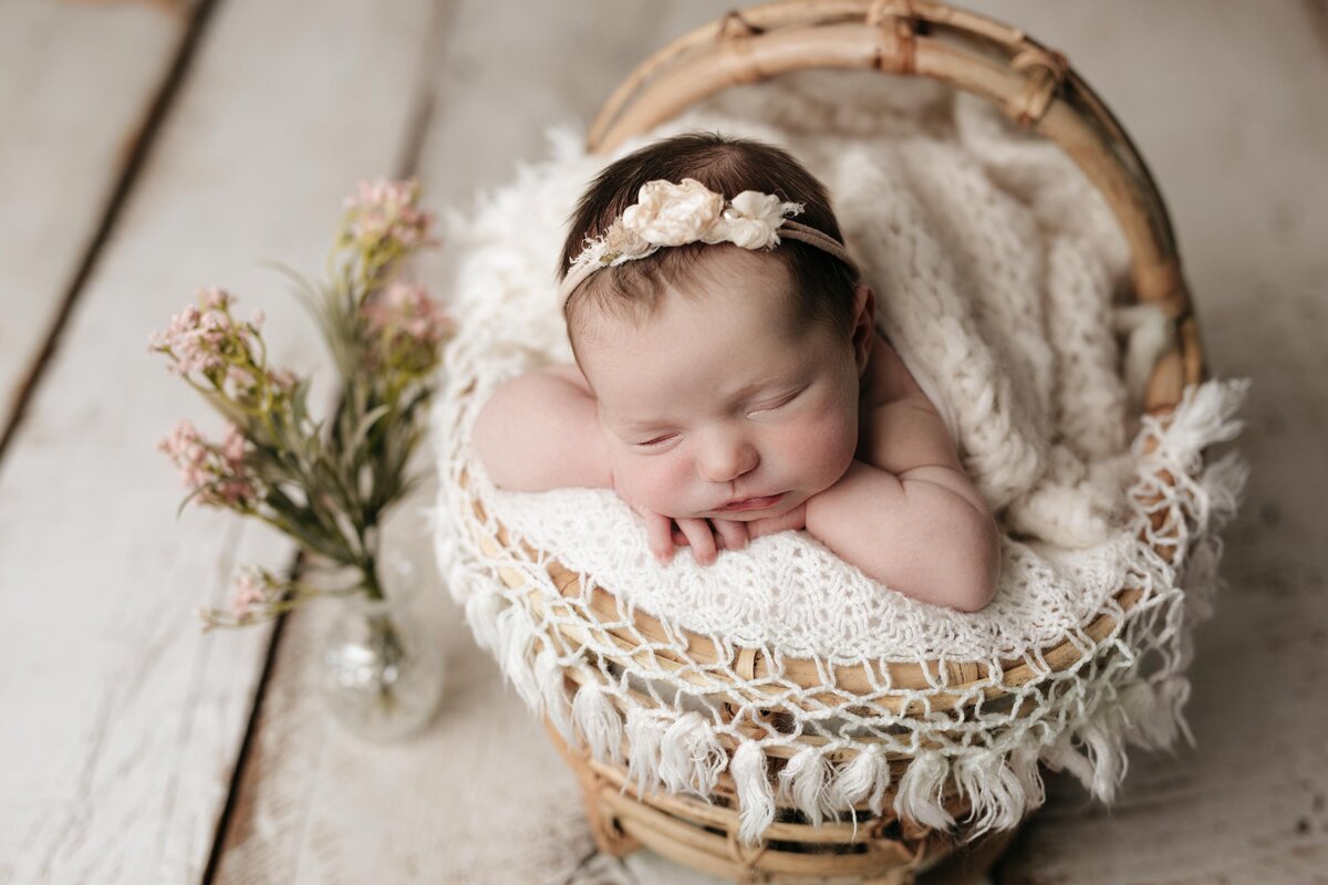 Studio Newborn Photography - Baby girl sleeping in a basket on a knit blanket with tassles that have a boho feel. Baby is wearing a floral headband. Her hands are resting under her chin.