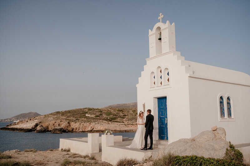 Couple échangeant ses vœux devant une chapelle blanche traditionnelle en bord de mer à Paros, en Grèce, lors d’un mariage à l’étranger intime et poétique.
