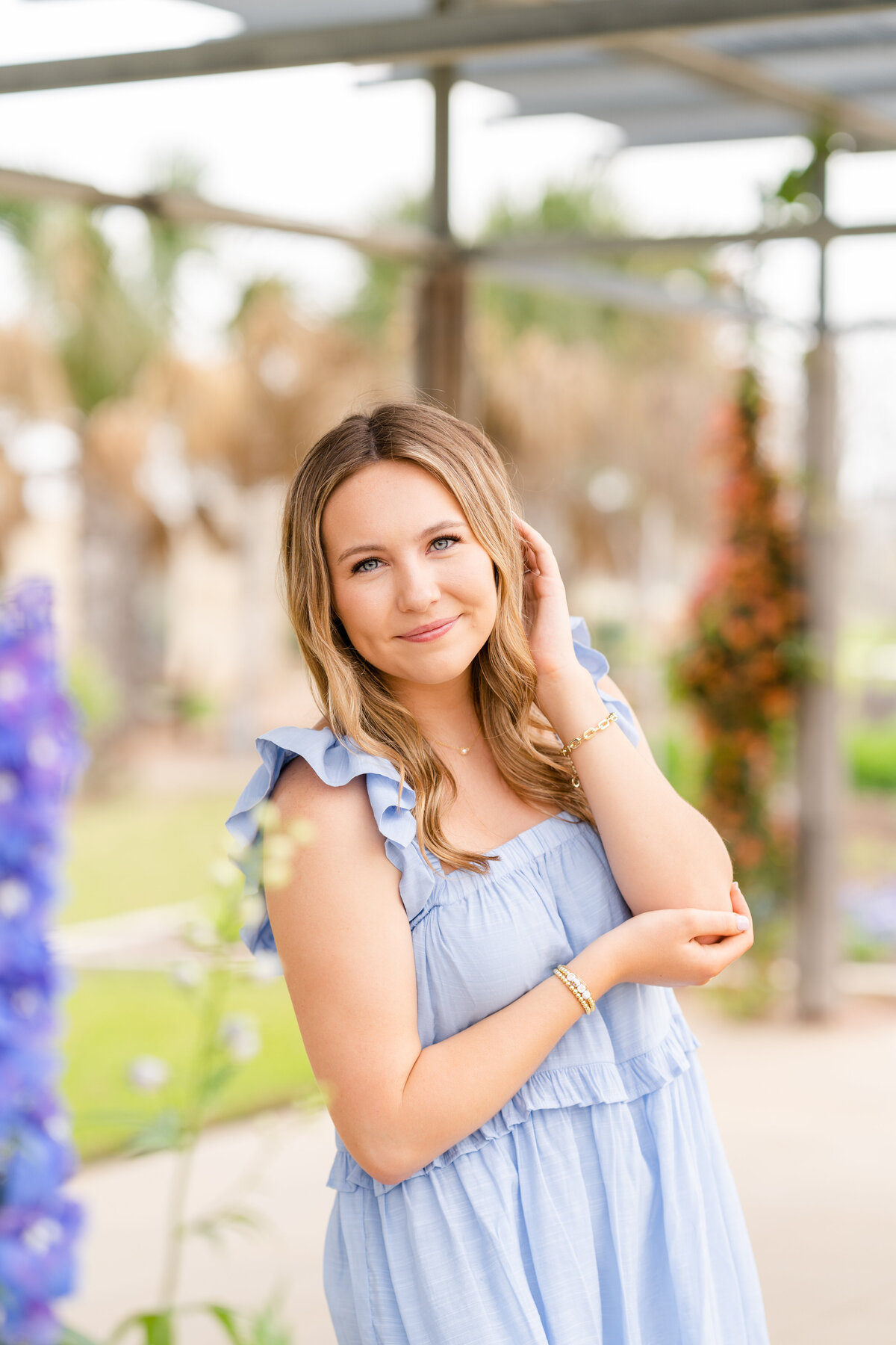 Texas A&M senior girl holding elbow and other hand in hair while wearing blue dress and surrounded by flowers at Leach Teaching Gardens