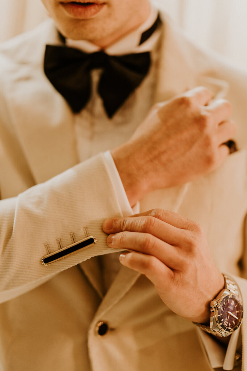 Homme en noeud papillon noir ajustant la manche de son costume beige agrémenté d'un bouton de manchette noir. Séance photo mariage.