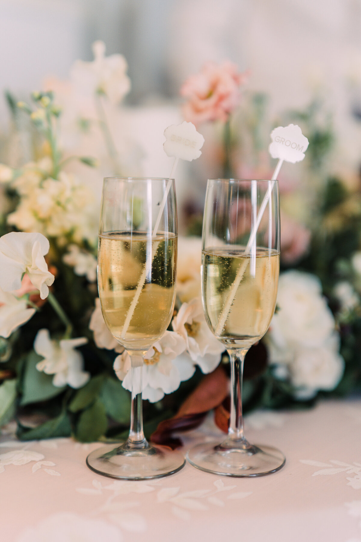 Two glasses of champagne with acrylic drink stirrers in front of white flowers, greenery, & burgundy foliage