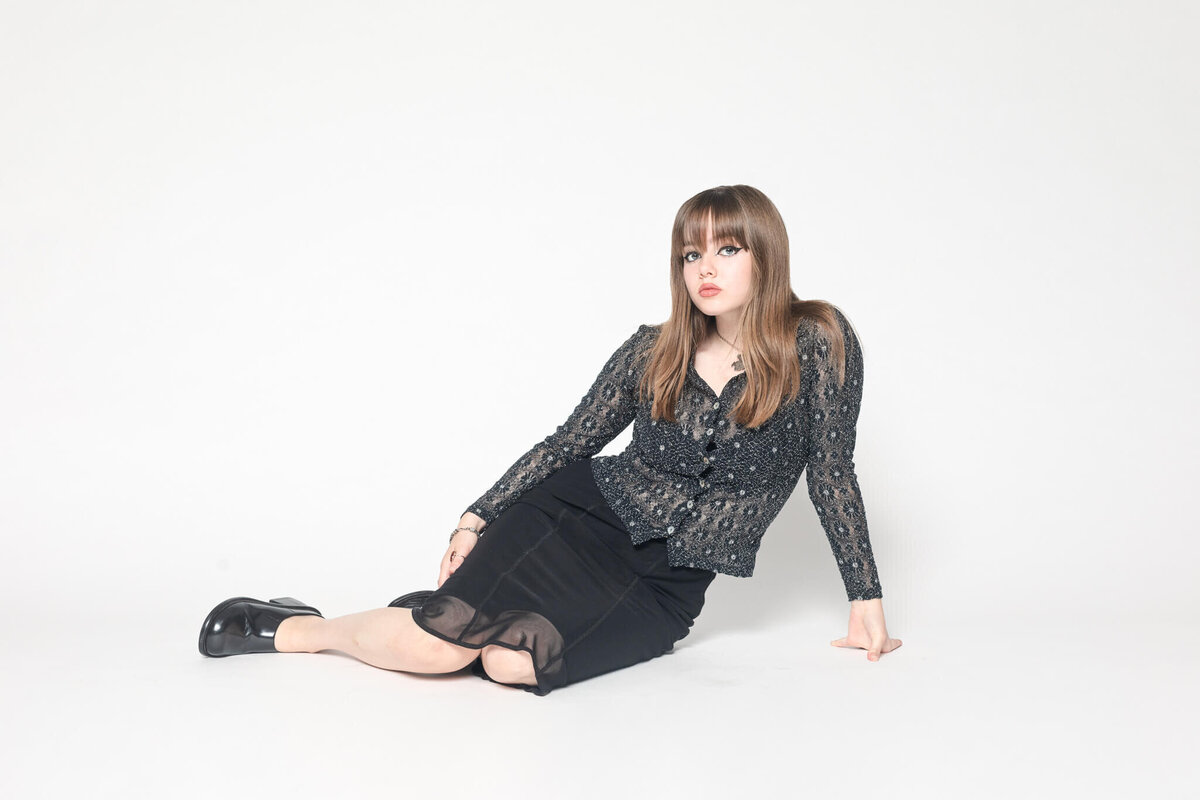 A person with long brown hair, wearing a patterned black shirt and a black skirt, sits on the floor against a white background. The individual is looking towards the camera with a relaxed posture.