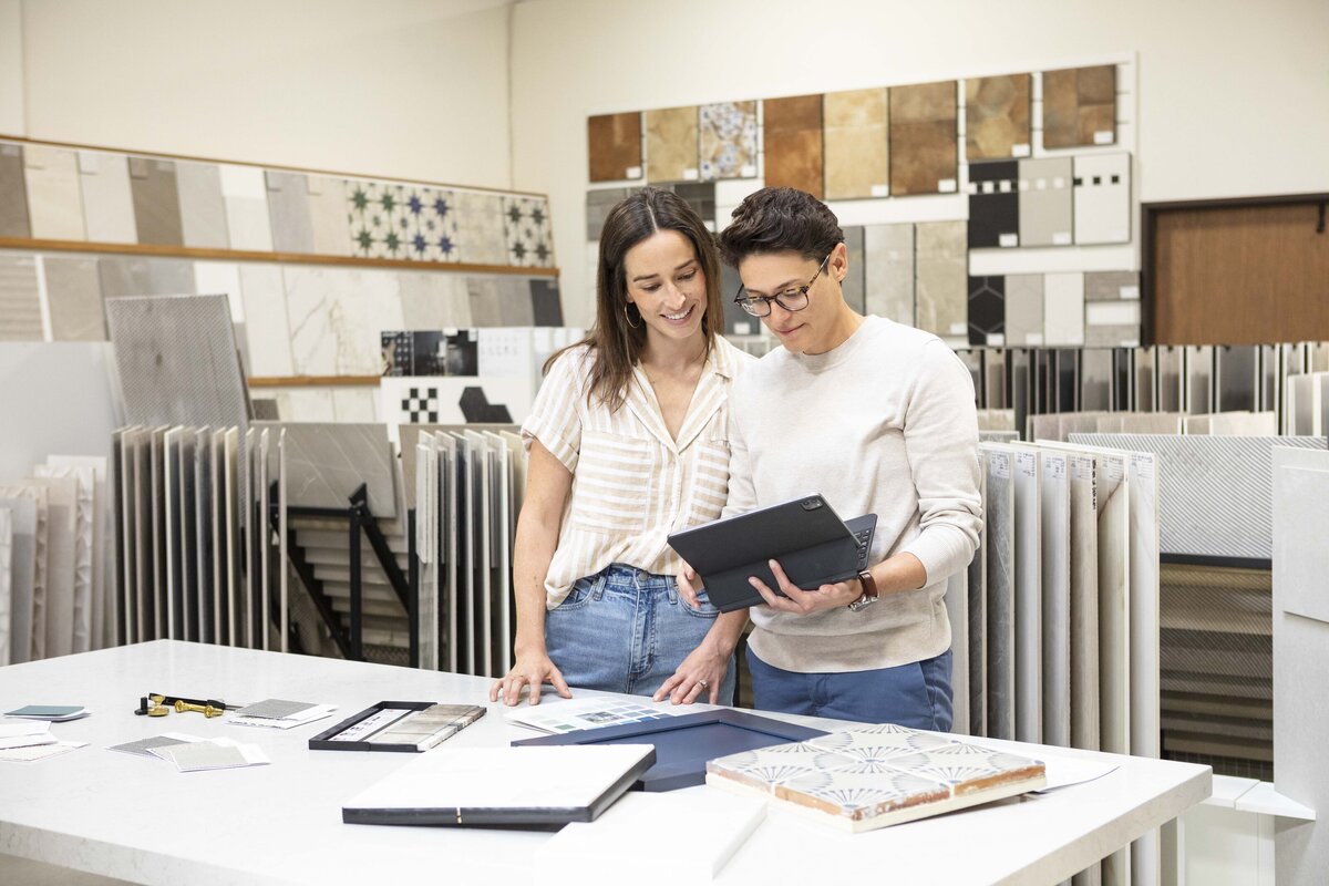 A designer duo reviews material samples and digital plans in a tile showroom, surrounded by large format tile displays and a worktable filled with design materials, at Tile City in Camarillo California.