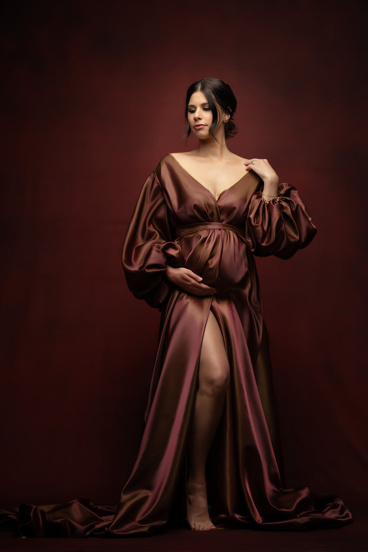 A pregnant woman in a flowing satin gown cradles her belly against a deep red backdrop. Captured by a Dallas maternity photographer, the portrait exudes elegance and confidence.