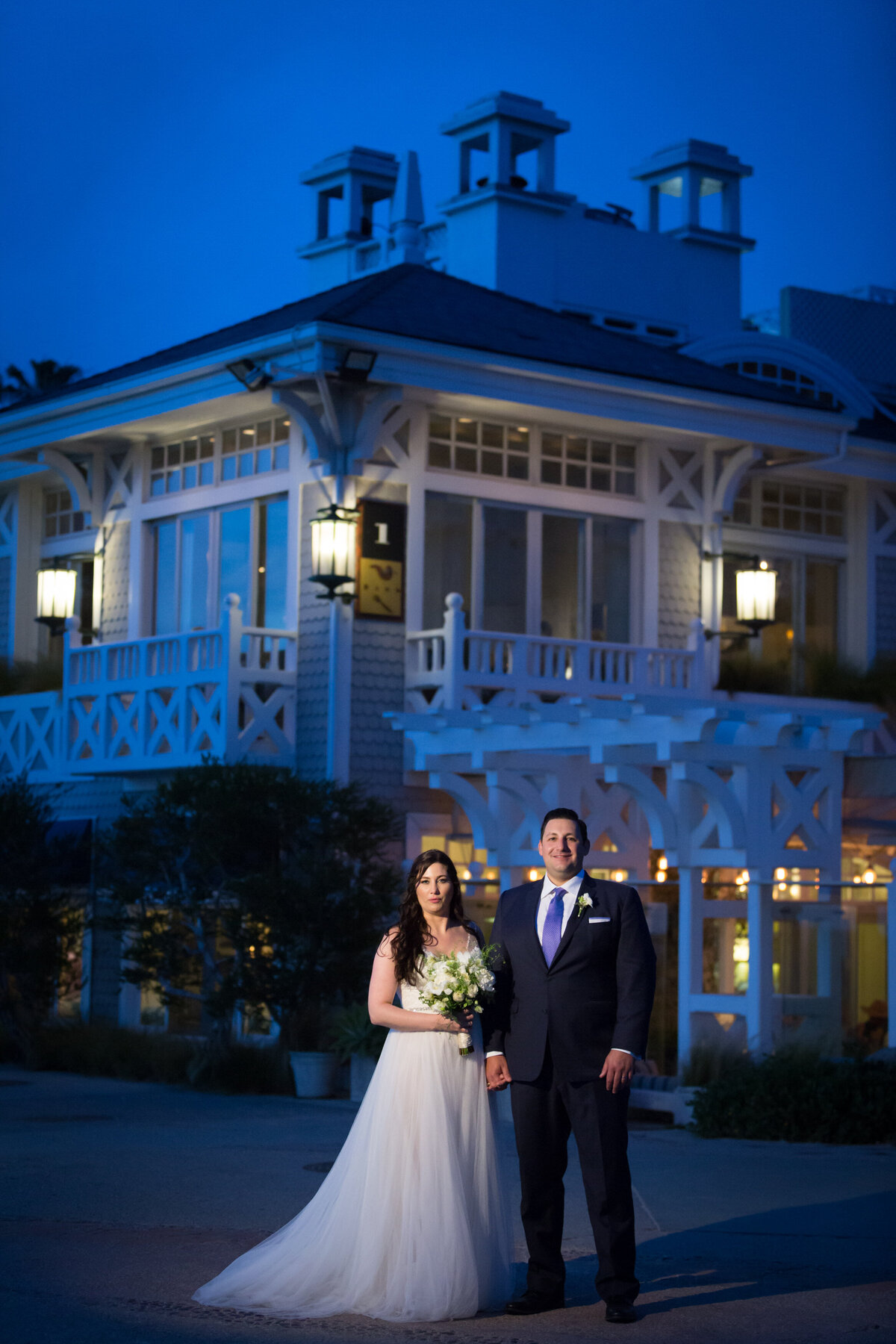 A newlywed couple holding hands while standing outside a large home