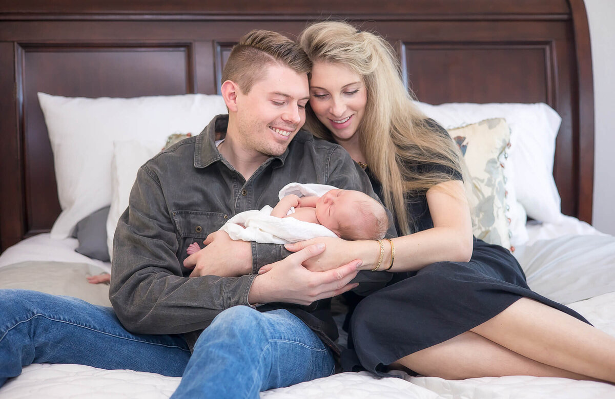 blissful mom and dad lovingly looking at newborn son and sitting on a bed