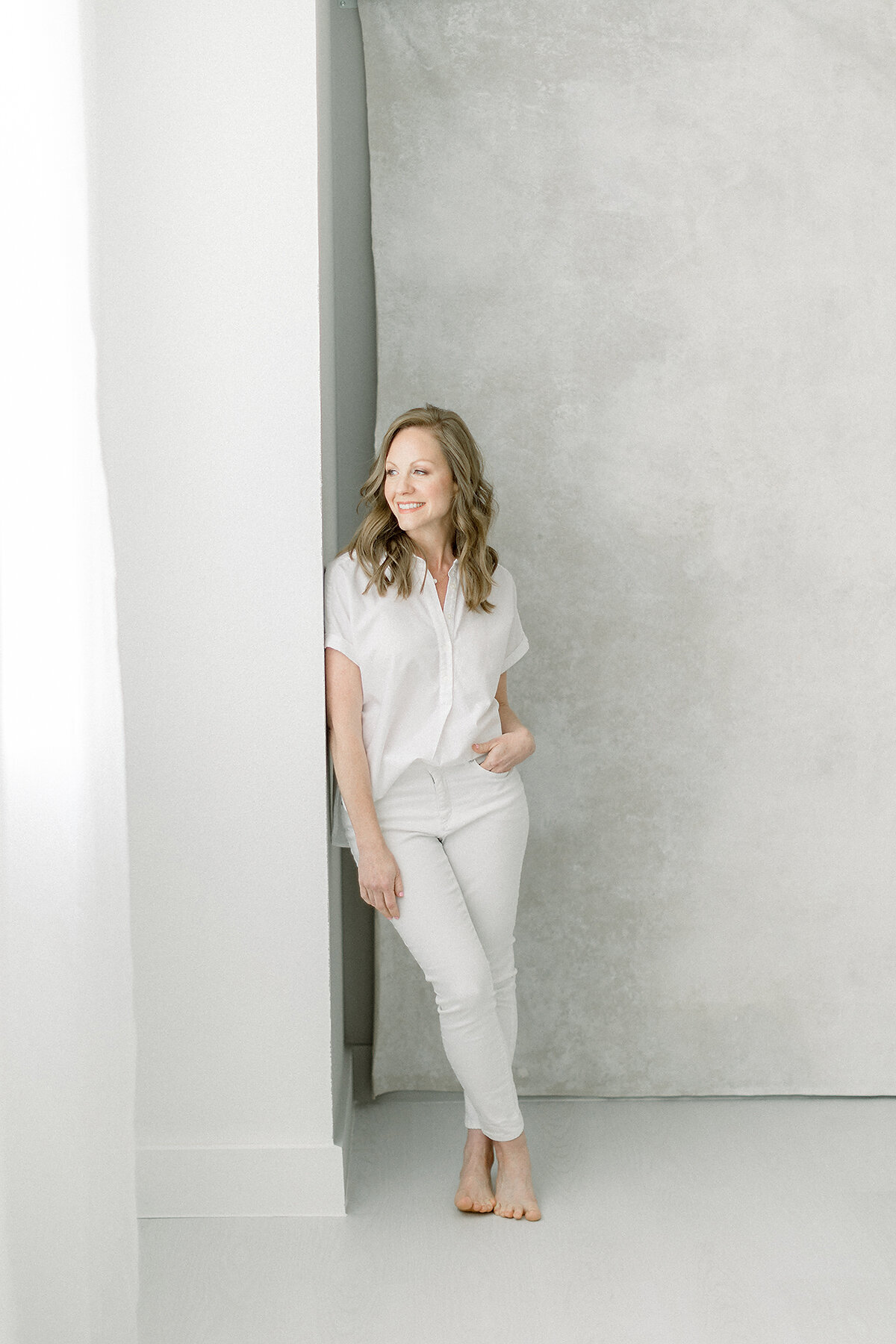 An in studio professional headshot taken of a Dallas business woman wearing a white shirt and pants as she looks out of the window and poses for her professional headshots.