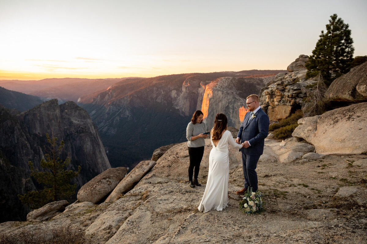 A couple stand holding hands and facing each other as their officiant reads from a book during their elopement ceremony in Yosemite.