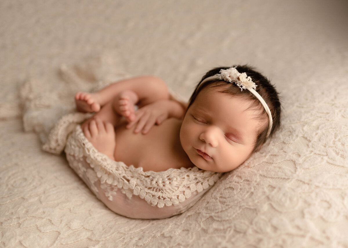Newborn girl sleeping curled up wrapped in lace on cream colored  lace with delicate floral headband