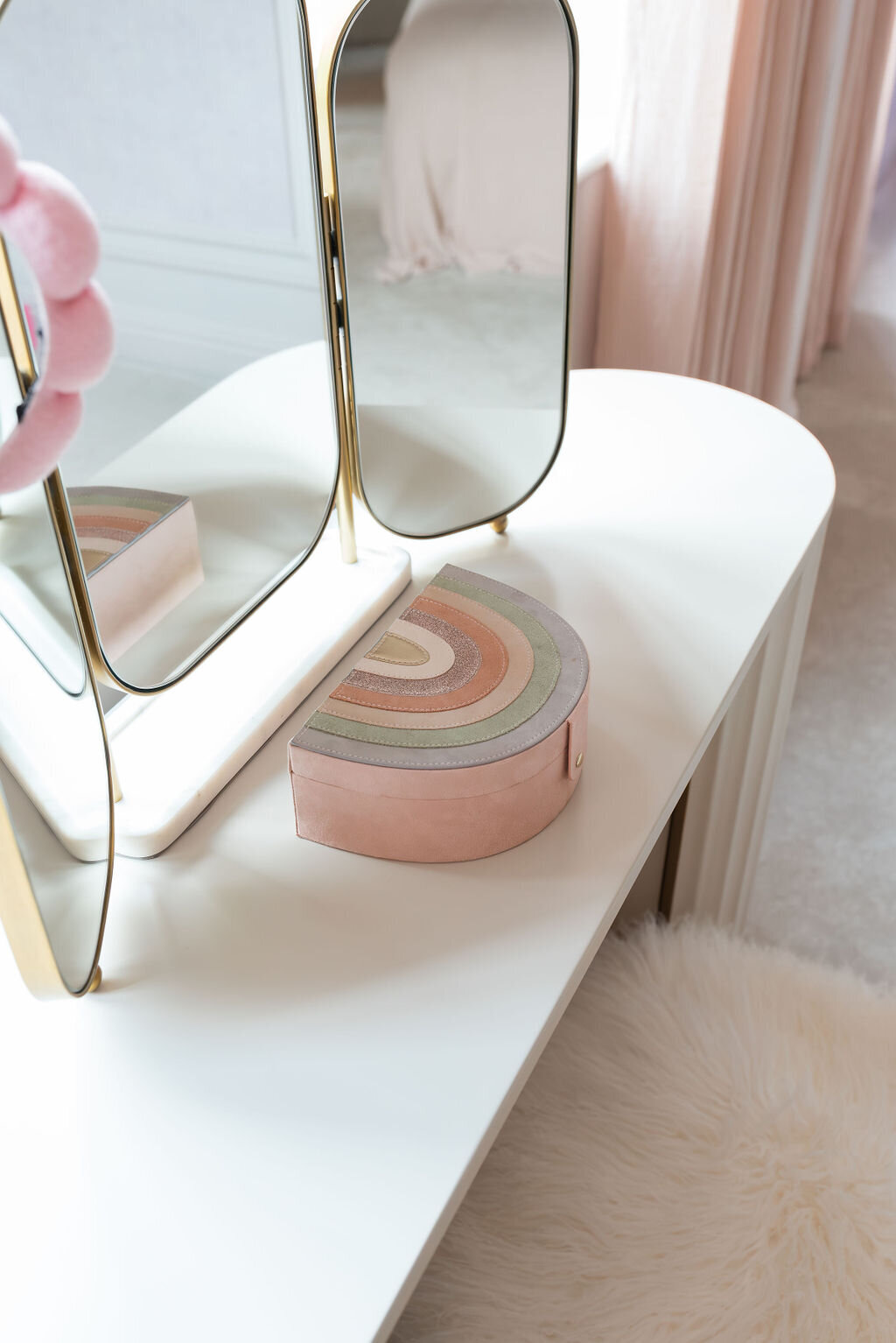 A white vanity table with a tri-fold mirror and a soft pink stool in a girl's pink bedroom.