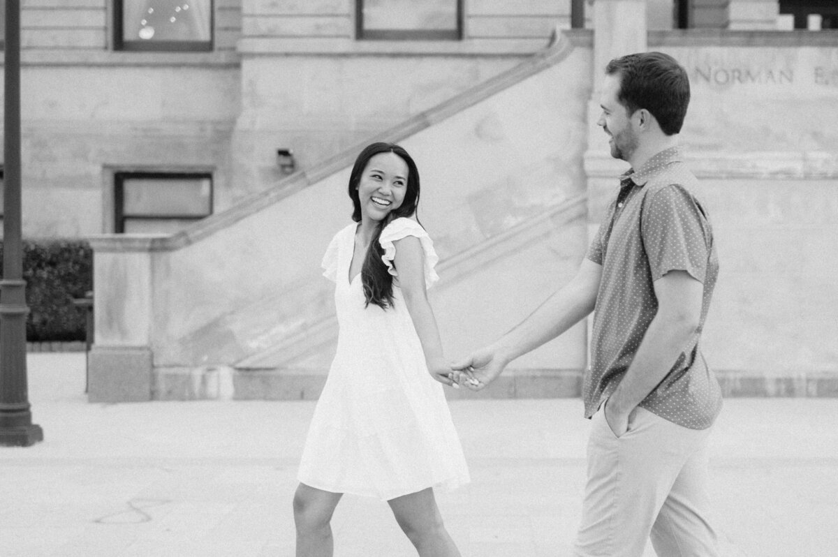 Black and white photo of a bride wearing a strapless dress with a bow and groom with a black tie tux walk hand in hand. Photo by Anna Brace, an Omaha wedding photographer.