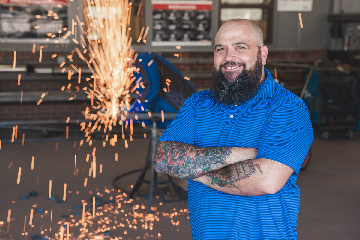 Male welding teacher with tattoos and a beard standing in front of sparks flying
