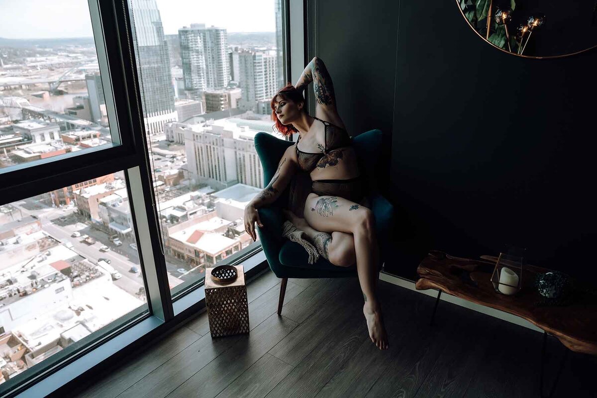 Tattooed woman posing on velvet armchair looking out of the window of a highrise Nashville apartment