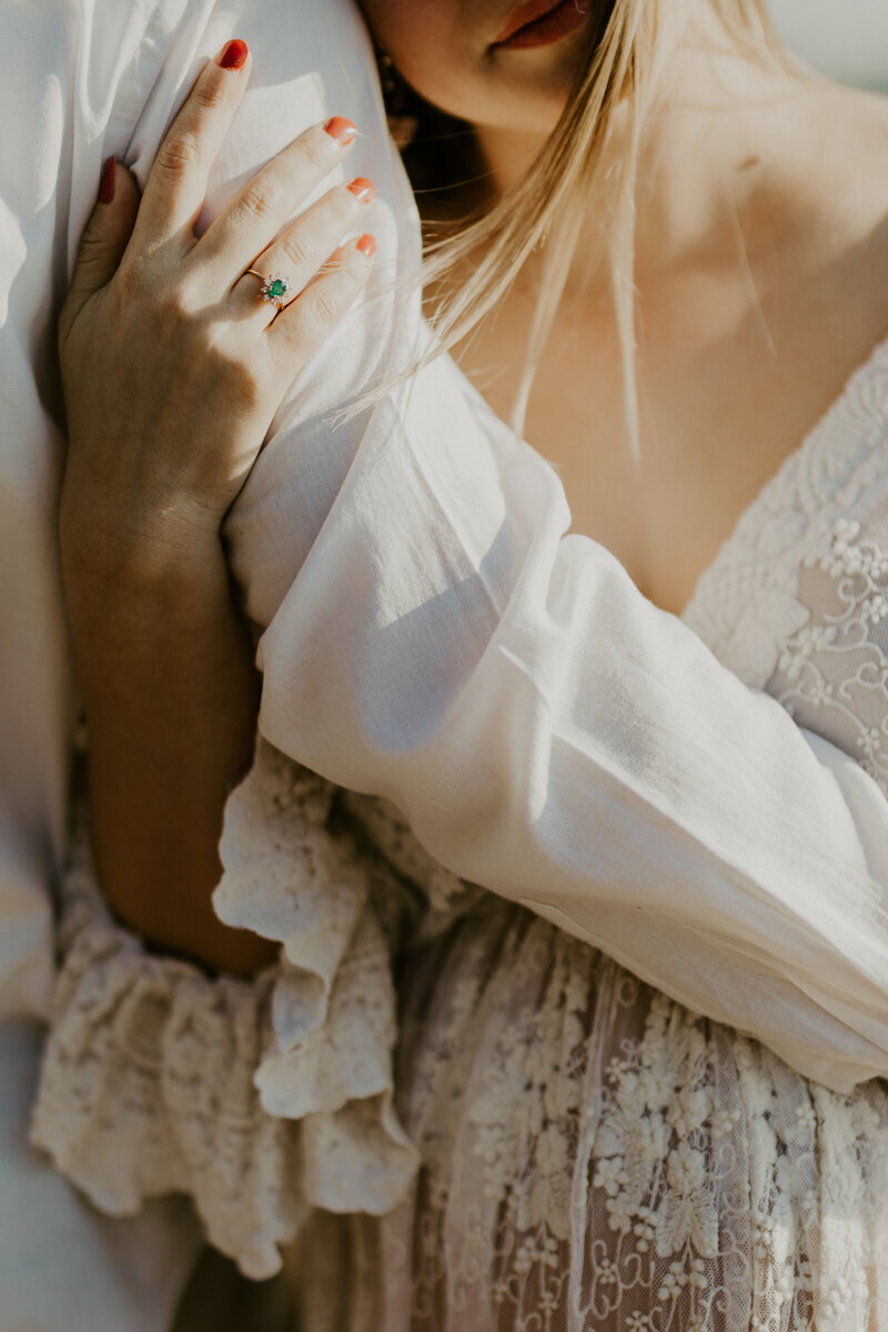 Zoom sur une main reposant sur le bras de son mari, enlaçant sa femme et son ventre rond recouvert d'une robe en dentelle lors d'une séance photo grossesse en vendée.