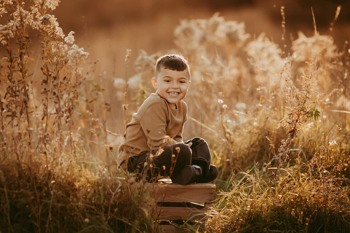 a little boy sitting criss cross  on a wooden crate in a beautiful golden field