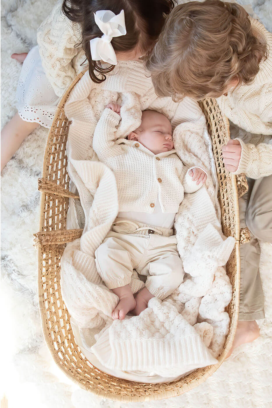 Two siblings look down at their newborn brother as he sleeps in a moses basket.