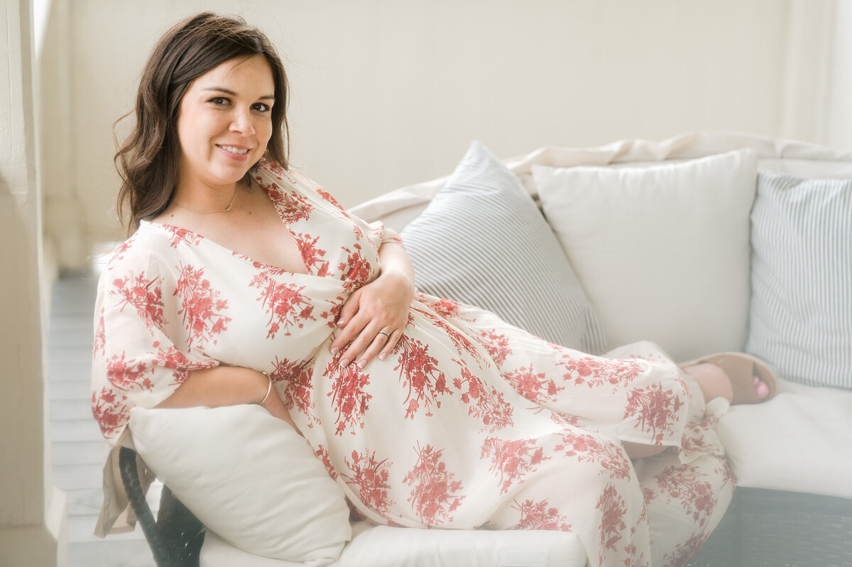 Expecting woman in a floral gown lounges on a couch and holds her bump.