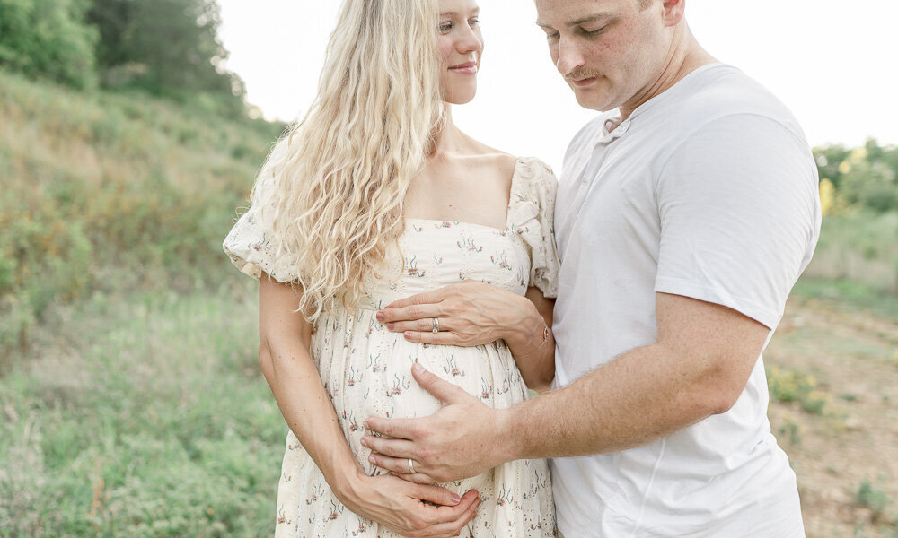 Pregnant woman smiles at her husband as he smiles at her stomach on a hill By Nashville maternity photographer Kristie Lloyd