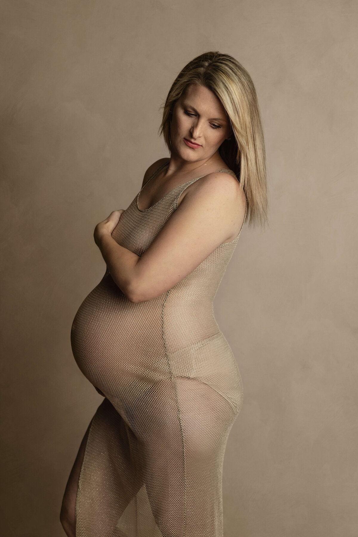 A blonde pregnant woman stands in a studio wearing a see through slip