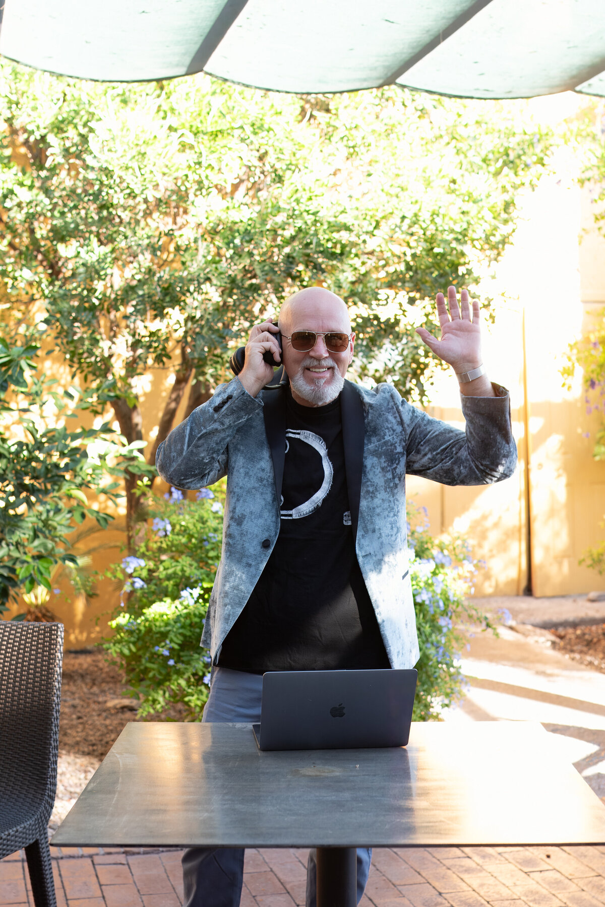 Man smiling while talking on the phone and waving outdoors, standing near a laptop with lush greenery behind him, photographed by Vyrl Photo for Tucson brand photography.