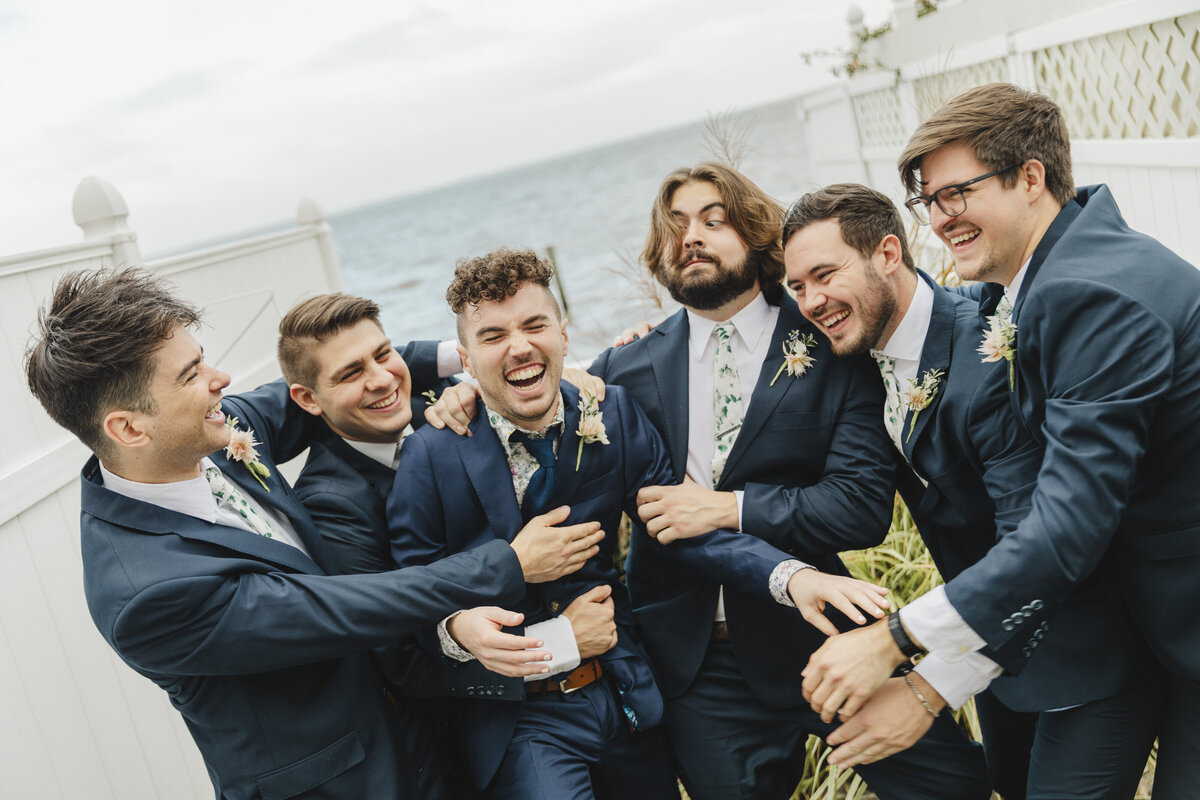 A wedding party tugging and hugging a newlywed as they all smile 