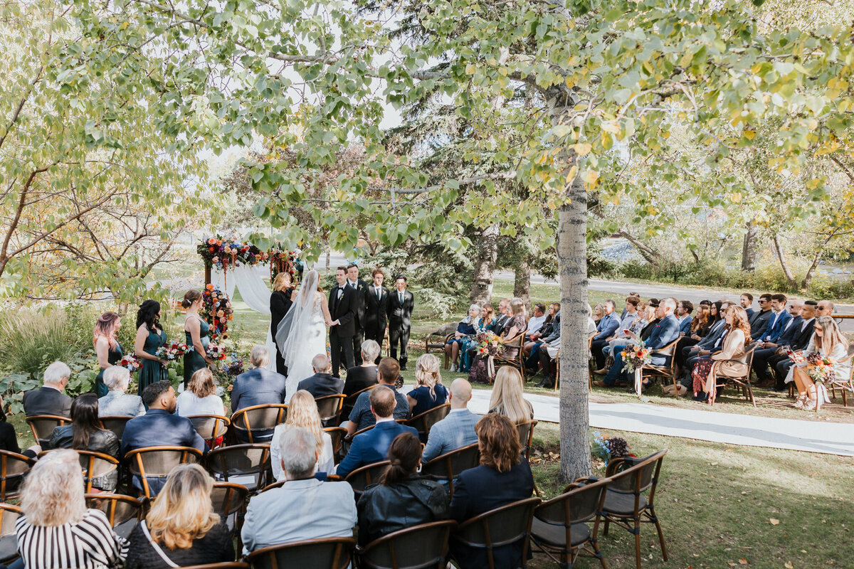 Outdoor wedding ceremony at River Café in Calgary, Alberta, featuring a couple exchanging vows under a vibrant floral arch. Surrounded by lush greenery, the bride in a flowing white gown and the groom in a classic black suit stand before their officiant. Bridesmaids in deep green dresses and groomsmen in black suits add to the elegant scene, while guests watch from floral-adorned wooden chairs. A romantic and picturesque setting along the Bow River.
