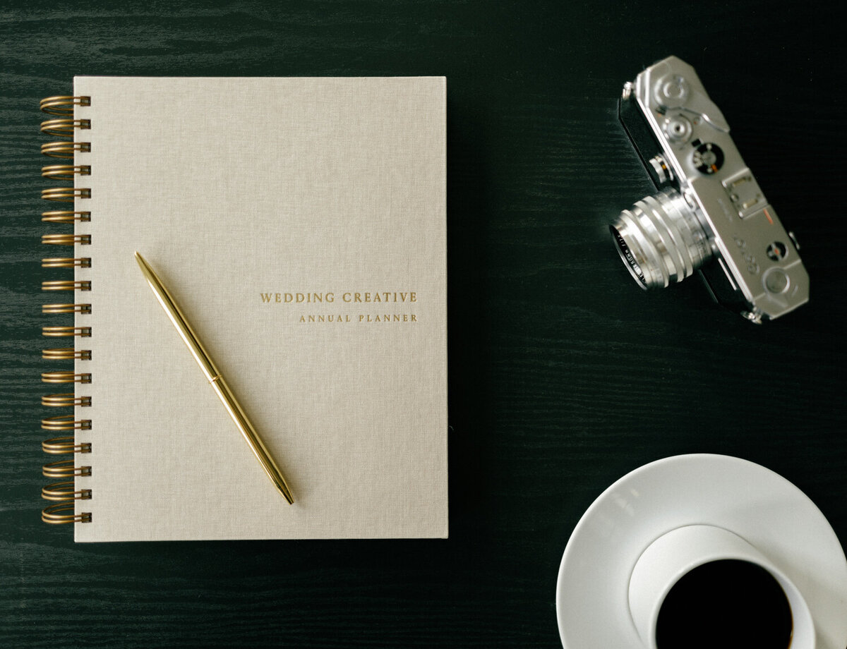 beige linen planner with gold lettering, coffee cup, vintage film camera on black table