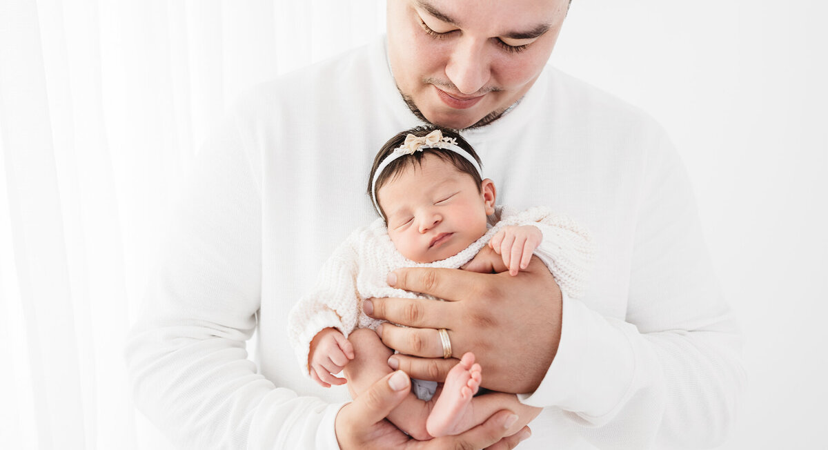 This heartwarming photograph captures a dad tenderly cradling his newborn baby girl. The father's gaze is adoring, filled with joy as he smiles down at the sleeping infant. The serene atmosphere is accentuated by the white backdrop, creating a clean and classic style. Shot by talented photographer Bri Sullivan.