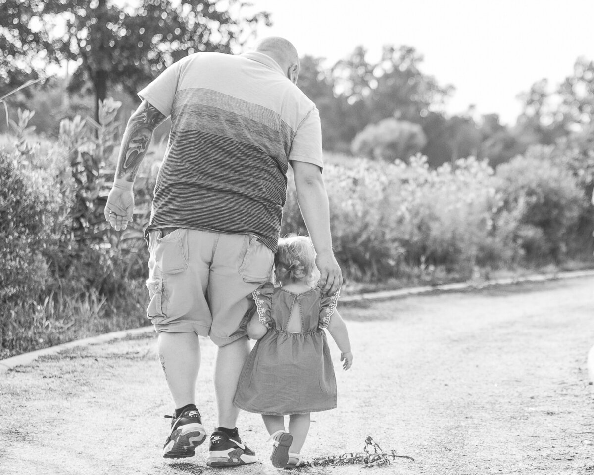 black and white photo of a dad walking with his toddler daughter.  Dad is wearing a stripped shirt and shorts looking down at his daughter.  Daughter is wearing a dress and walking next to dad