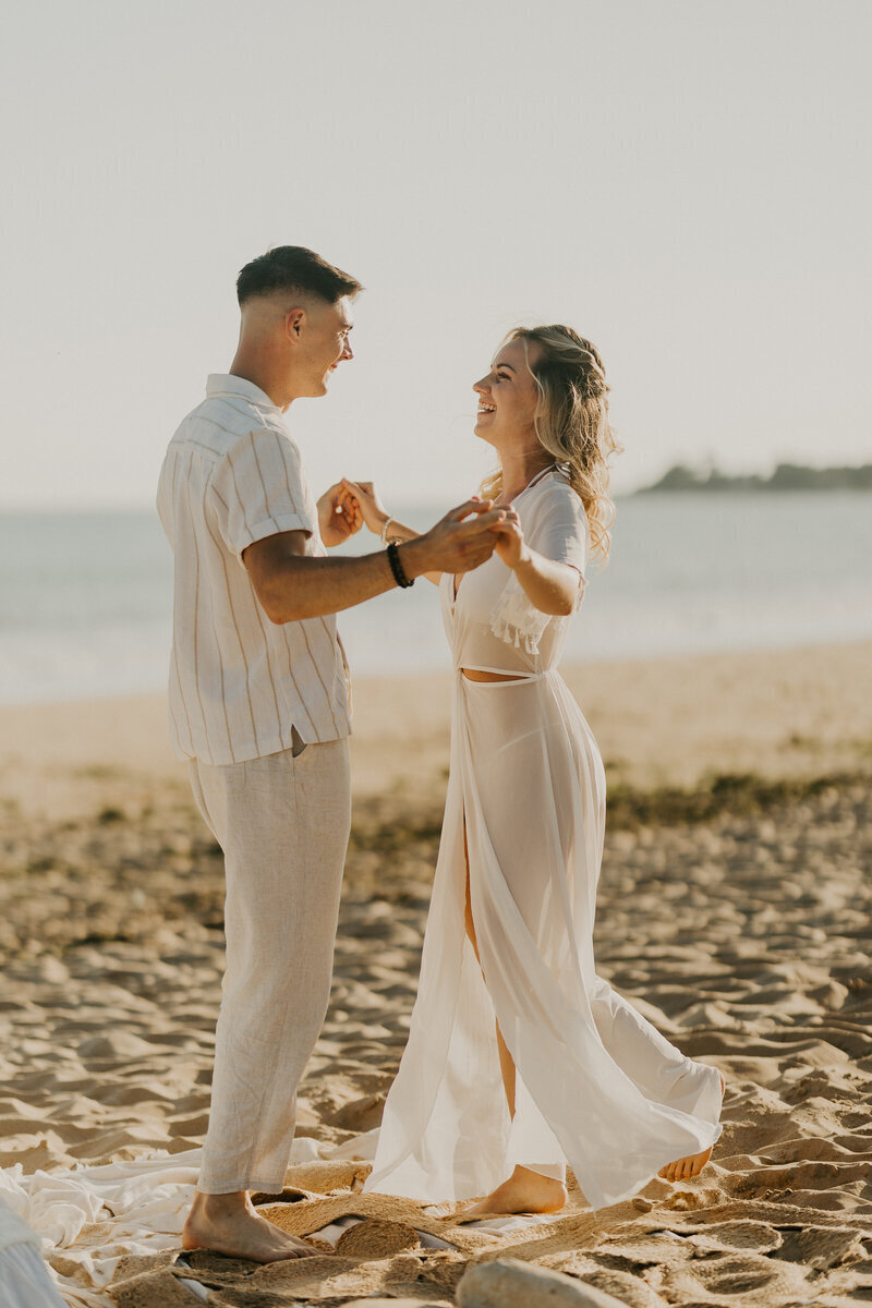 Couple en tenue blanche, mains jointes, dansant ensemble dans un décor de plage lors d'un shooting photo couple.