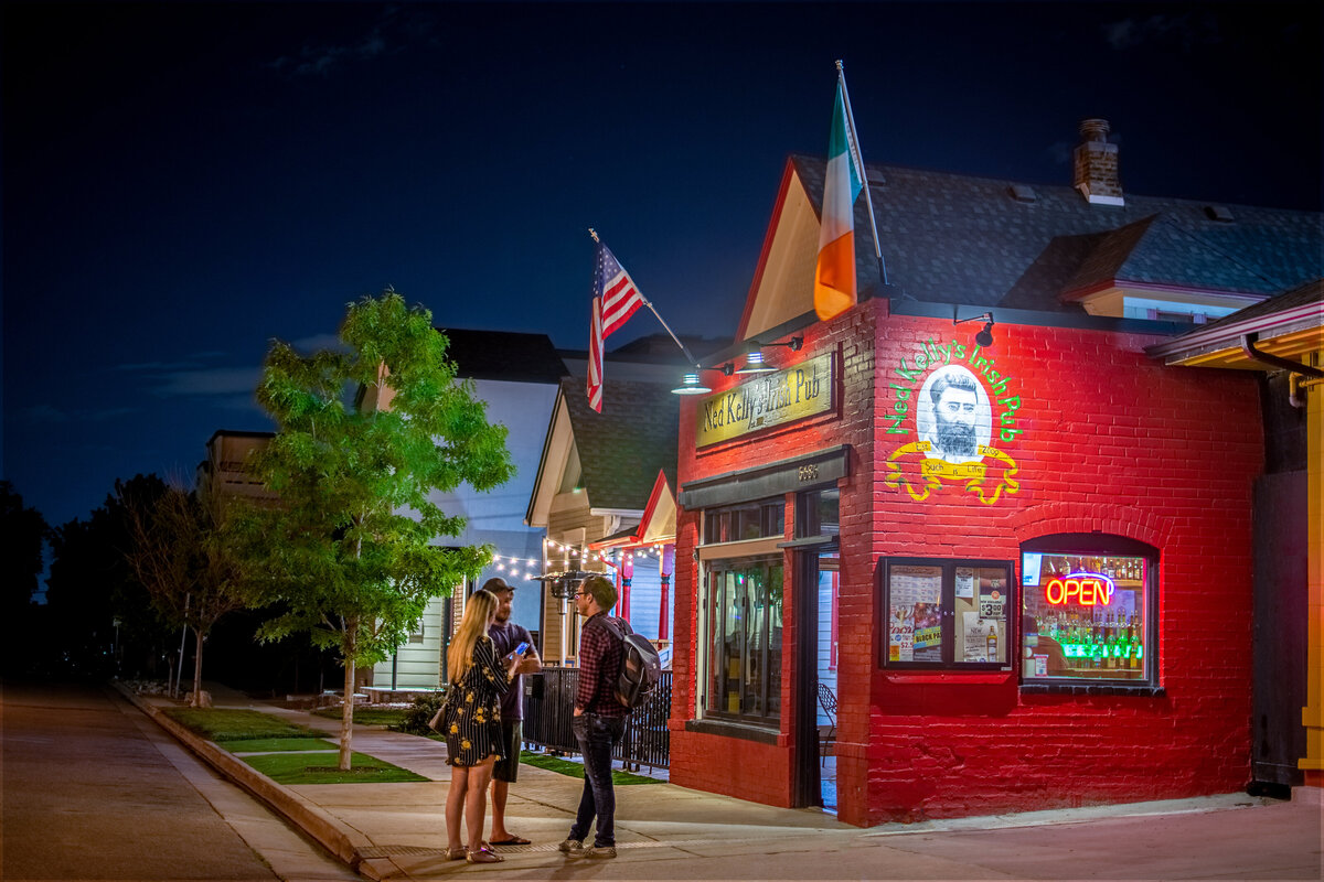 A group of three young adults stands on the sidewalk outside of Ned Kelly's Irish Pub in Downtown Littleton on a summer night.