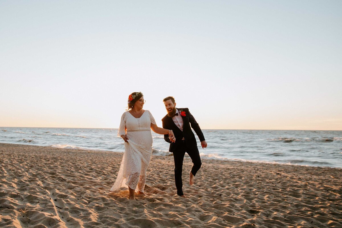 Bride and groom walking along Grand Bend beach at sunset for top wedding portraits. They are holding hands and looking at each other with warm golden pink light.