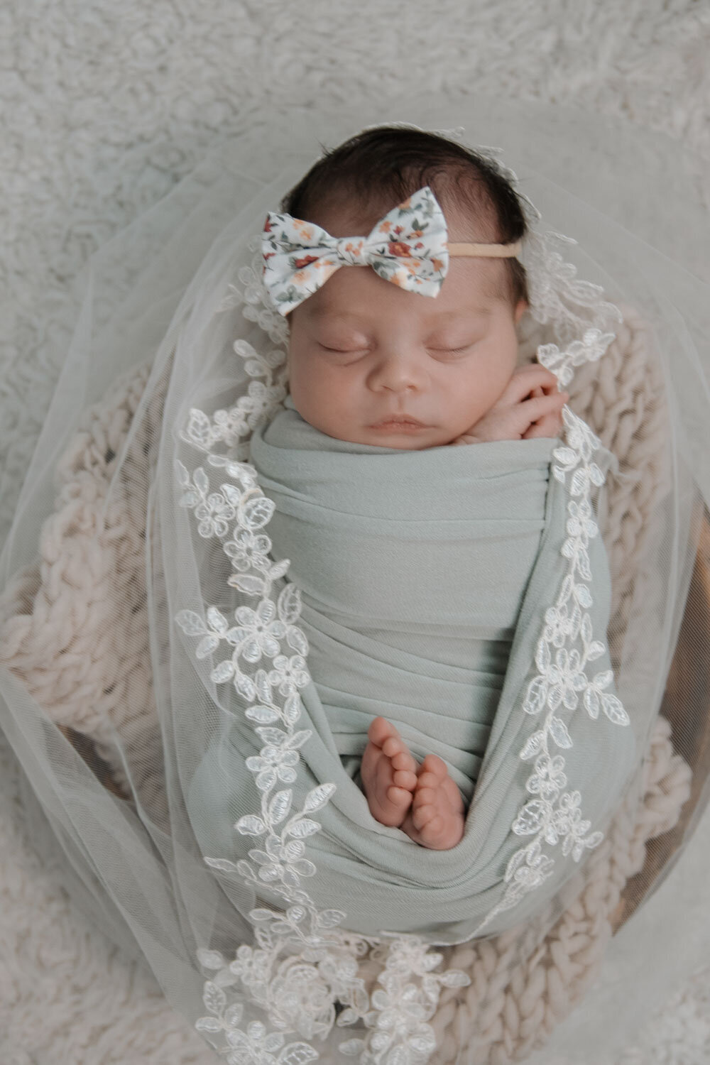 Baby wrapped in a green swaddle with lace accents, wearing a floral bow headband, lying peacefully