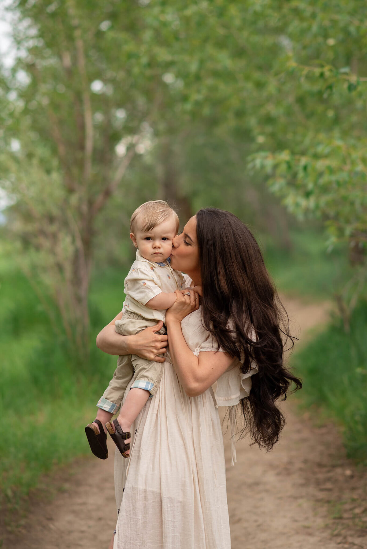 A mother holds her baby boy and gives him a kiss on the cheek. Taken in a beautiful park in Kamloops and captured by Brittany Danielle.